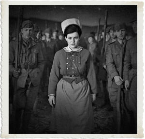 A serious young woman in a World War I-era military nurse uniform stands front and center at attention, gazing directly forward with a focused expression. She wears a white nurse’s cap, dark jacket with white sailor-style collar, and belt. She is surrounded by rows of male soldiers in uniform, all standing rigidly at attention while holding rifles vertically in a large, dimly lit military barracks or tent. The image is in dramatic black-and-white with a vintage aged photo border.