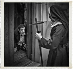 In a dramatic black-and-white scene, a terrified man in a tuxedo with vampire fangs kneels in a doorway, mouth open in a shout or hiss, as a nun in traditional habit stands over him holding a large wooden cross like a weapon. The nun faces him with determination while the man cowers on the floor. The image has a vintage, aged photo border and a cinematic horror style.