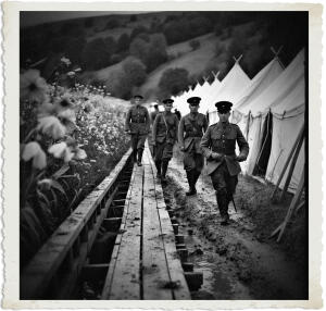 Black and white photograph of four soldiers in early 20th-century British-style military uniforms walking toward the viewer along a muddy path beside a wooden boardwalk. White flowers bloom prominently in the foreground on the left. In the background, a long row of canvas military tents lines the path under a dark, overcast sky with rolling hills.