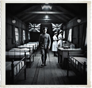 Black and white photograph of a World War I field hospital ward inside a long wooden hut. A young British soldier in uniform walks confidently down the wide central aisle toward the viewer, holding a walking stick. Metal beds are lined along both sides of the room. In the background, two Union Jack flags hang on the far wall. Two nurses in white uniforms work at a desk on the right side of the room.