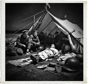 Picture of an aid station near the front tend, white tent is damaged and dirt, bandages, blood, helmet and gear lying around. Medical staff look exhausted. Clothing and style appropriate for 1915 for No Man Land at night.