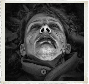 Black and white close-up photograph from the “Man at War – Carried by Six” series. A young soldier lies on his back with his head tilted upward, eyes open but vacant. His face is covered in dirt and grime, and his lips have a dark, bluish tint. The image is intimate and haunting, conveying death, exhaustion, or the final moments of life on the battlefield. A vintage distressed border frames the somber portrait.