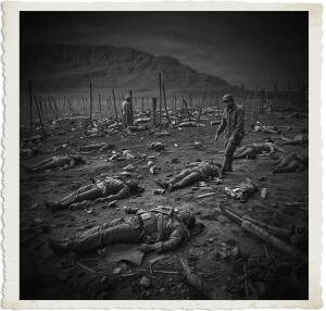 Black and white photograph from the “Man at War – Carried by Six” series. A devastated battlefield is strewn with dozens of fallen soldiers lying motionless across the muddy ground amid scattered equipment and wooden stakes. In the right midground, a lone helmeted soldier stands grimly surveying the carnage, while another figure stands farther back. The barren, mountainous landscape and heavy sky create an overwhelming sense of slaughter and desolation, evoking the horrific cost of war.