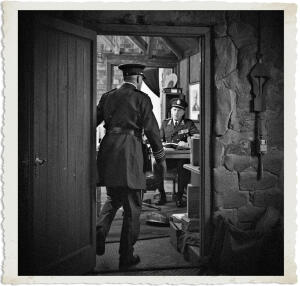 Viewed from behind, a senior officer in a dark military greatcoat and peaked cap walks through an open wooden doorway into a stone-walled office. Inside, a younger officer seated at a desk looks up toward him with a neutral expression. The room contains a desk with papers, chairs, framed pictures, and military equipment. The entire scene is captured in dramatic black-and-white with a vintage aged photo border.