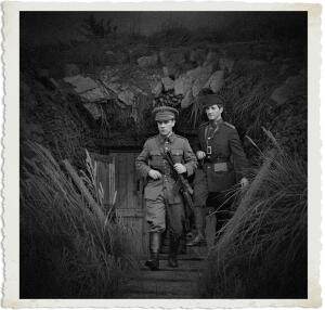 Two British officers emerge from a stone and wooden bunker entrance set into a rocky, grassy hillside. The officer in front, wearing a peaked cap and service dress uniform, holds a rifle and looks forward with a serious expression. The second officer, in a darker uniform with a side cap, stands slightly behind him. Tall grass and reeds frame the scene in the foreground. The image is in dramatic black-and-white with a vintage aged photo border.