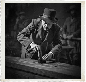 Black and white photograph of a somber man in a fedora and heavy overcoat leaning over a wooden coffin, driving a nail into the lid with a hammer. His gaze is focused downward in quiet concentration. In the softly blurred background, other uniformed figures stand nearby.