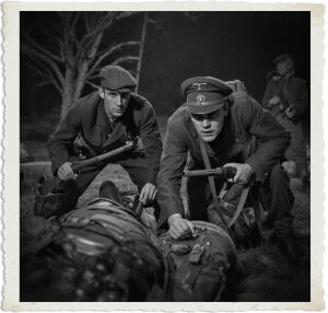 Black and white nighttime photograph from the “Man at War – Carried by Six” series. Two soldiers in dark WWII-style uniforms crouch tensely in the grass. The soldier on the right, wearing a peaked officer’s cap, grips a submachine gun and stares forward with focused intensity. The soldier on the left, in a flat cap, holds a rifle and looks ahead with a grim expression. Military gear and packs lie in the foreground while a third figure is faintly visible in the shadowed background. The scene is moody and cinematic, evoking a tense night patrol or ambush.