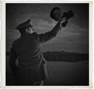 Black and white photograph of a man in a military-style coat and peaked cap standing with his back partially to the viewer. He holds aloft a severed human head (still wearing a soft cap) in his outstretched right hand, silhouetted against a dark, brooding sky. A distant lighthouse glows faintly on the horizon over a body of water.