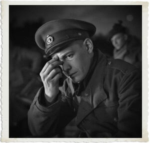 Black and white close-up portrait from the “Man at War – Carried by Six” series. A young officer in a military cap and uniform looks down and to the side with a weary, pained expression. He raises one hand to rub the bridge of his nose or wipe his eye, as if fighting back tears or exhaustion. Another soldier is softly blurred in the background. The image conveys deep fatigue, sorrow, or emotional strain amid the hardships of war.