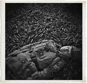 Black and white photograph of a young soldier in full military uniform and helmet lying on his back atop a vast, dense pile of thousands of spent shell casings that stretch into the background. His eyes are open, gazing upward, with a calm yet somber expression. The image has a vintage, distressed border and evokes the heavy toll of war.
