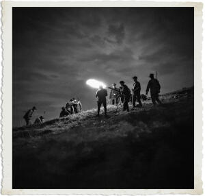 Black and white nighttime photograph from the “Man at War – Carried by Six” series. A group of soldiers stands on a grassy hill under a dramatic, cloudy sky. One soldier in the center holds up a bright flare or spotlight that illuminates the scene, while others stand watch with rifles. Some are crouched or operating equipment on tripods. The composition has a tense, cinematic feel, suggesting a night patrol, artillery spotting team, or forward support unit preparing for action.