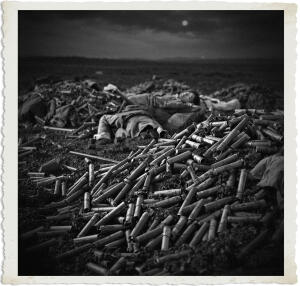 Black and white nighttime photograph from the “Man at War – Carried by Six” series. A soldier lies motionless on his back atop an enormous mound of thousands of spent shell casings that blankets the foreground of a barren battlefield. Other fallen soldiers are scattered among the massive pile of brass and debris under a dark, cloudy sky with a visible moon, powerfully conveying the staggering scale of firepower and the overwhelming human cost of war.