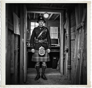 British soldier inside of a bunker. He wears a kilt and a brodie helmet stands at the treshold to an office. Background showing sings of this being a headquarters. Setting and clothing appropriate for 1914 in No Mans Land.