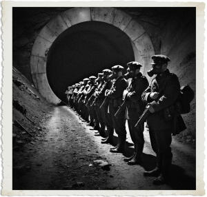 a surreal and vibrant cinematic photo of British soldiers lined up by a large tunnel entrance, heavily armed with rifles and pistols, and wearing gas masks, their clothing and style appropriate for 1915, set in No Man's Land at night, with a dark and ominous atmosphere, the soldiers' faces obscured by the masks, their eyes glowing in the dark, the tunnel behind them a deep and foreboding black, the scene captured in a photorealistic style with film grain, the colors muted and subdued, with hints of blue and green, the overall mood tense and foreboding, the soldiers' bodies silhouetted against the faint moonlight, their shadows cast long and sinister on the ground.
