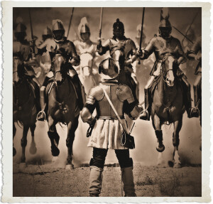 French soldier in late 16th-century armour stands with his back to the viewer, facing a charge of armoured cavalrymen at full gallop. Dust rises under the horses’ hooves on an open field under bright daylight. Sepia-toned period photograph.