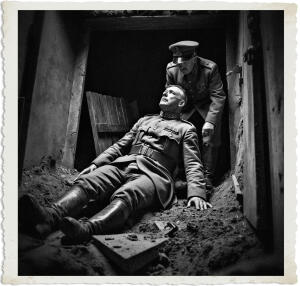 Black-and-white vintage-style photograph with a torn-paper border, showing a dramatic wartime scene inside a damaged bunker or trench. A senior military officer in full uniform and peaked cap lies collapsed on the dirt floor, head tilted back, while another high-ranking officer stands over him, looking down with concern amid rubble and debris.