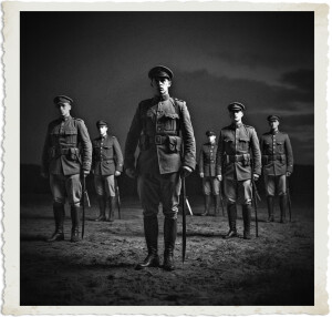 Black and white vintage-style photograph of soldiers standing at attention in a barren field at dusk. A stern young officer stands prominently in the foreground holding a rifle, with four other soldiers positioned behind him in formation. The dramatic, moody lighting and aged photo border evoke early 20th-century military discipline.