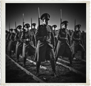 Black and white vintage-style photograph of soldiers performing drill in formation on a field at night. A stern officer stands prominently in the foreground holding a rifle with bayonet, with other soldiers marching in line behind him under dramatic lighting. The image has a classic aged photo border.