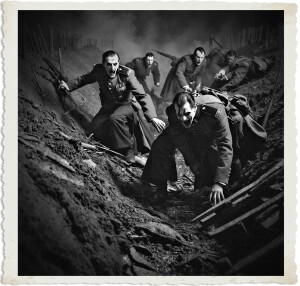 Black and white dramatic photograph of vampiric soldiers with deathly pale, contorted faces charging aggressively out of a muddy trench. In the foreground, one officer brandishes a submachine gun while others scramble forward with open-mouthed screams. The chaotic nighttime battlefield scene features smoke and debris, styled as a vintage photograph with torn-edge borders.