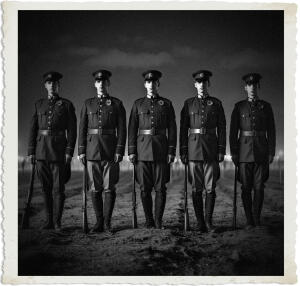 Black-and-white vintage-style photograph with a torn-paper border, showing five soldiers in early 20th-century military uniforms standing in a formal line on a muddy field. Each holds a rifle at their side and wears a peaked cap with a poppy pinned to the chest, gazing directly forward under a dark, somber sky.