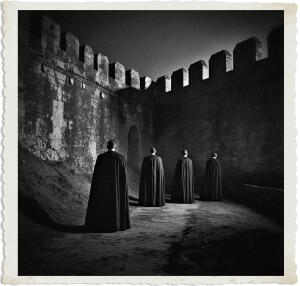 Black and white dramatic photograph of four men wearing long dark capes standing with their backs to the viewer, facing a towering ancient stone fortress wall at night. The men are lined up in a row, looking toward the high battlemented walls under dramatic low-key lighting that creates strong shadows on the ground. The image has a vintage aged white border.