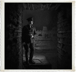 Black and white atmospheric photograph of a uniformed military officer standing in the shadows inside a dark, ruined stone fort, leaning against a wall and gazing into a dimly lit passageway.