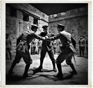 Black and white dramatic photograph of three British Army officers in a tense standoff inside a stone fortress courtyard at night. Two officers in peaked caps and uniforms aggressively grab each other’s arms while facing off, as a third senior officer with medals stands between them, watching intently. Other soldiers observe from the background. The image has a vintage aged white border.