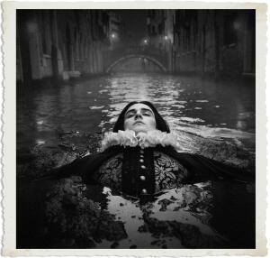 Black and white dramatic photograph of a pale figure with long dark hair floating motionless in a Venetian canal at night, only their head, shoulders, and upper torso visible above the dark water. The figure wears elaborate 17th-century noble attire featuring a large white lace ruff collar and ornate dark doublet. In the background, misty canal buildings and a stone bridge are visible under low lighting. The image has a vintage aged white border.