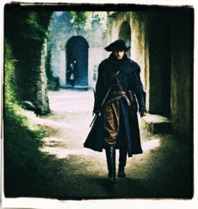 An assassin creeps out from out of the shadows with a dagger in hand. Approaches an unsuspected and unarmed teenage noble. Clothes and background appropriate for 16th century France.