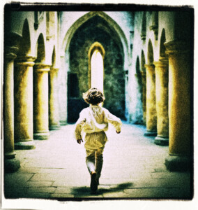 Picture of a young boy running away fromt he acmera. Appears to be running through a castle with several columns visible and arched doorways.
