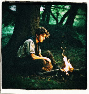 18 year old man in the woods sitting against a large tree. Man is looking over a campfire with something cooking atop the flame. Clothes and background appropriate for 16th century France.