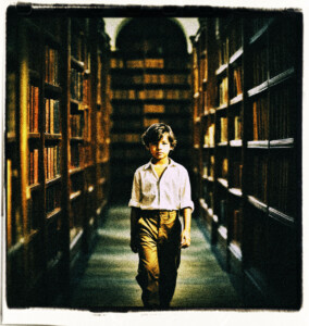 Picture of a boy with brown hair, black eyes, and a white shirt, walking through a library, with books towering over him.