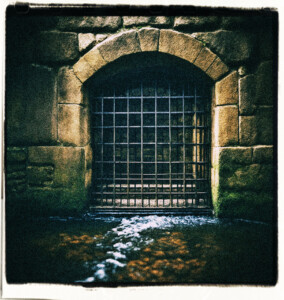 Picture of a grate with heavy iron vertical bars, on the floor or against the wall, the other side is dark, with water running through. Clothes and background appropriate for 16th century France.