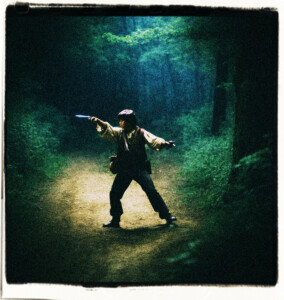 Teenage boy pointing his pistol at a man wielding a sword. Located in the dark woods at night, near a dusty road. Scene and clothes appropriate for 16th century France.