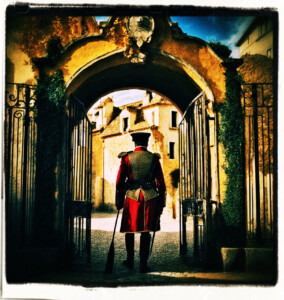 Guard exits the gates to deliver a message, leaving another guard behind standing at attention. Background and scene appropriate to 16th century France.