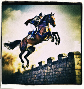 Picture of a horse and rider jumping over a stone wall that could belong to a castle. Dust behind the horse implies they were traveling at great speed.