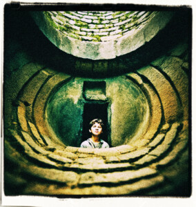 Teenage boy looking up from inside of a deep well, barely any light filters down. Clothes and background appropriate for 16th century France.