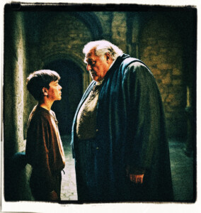 The picture of a corpulent old man facing off a teenage boy in dark castle halls. The man appears dark and foreboding as though from a nightmare. Clothes and background appropriate for 16th century France.