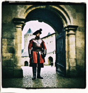 Two armed guards standing at the castle gates. One elbows the other for something he said. Background and clothing appropriate to 16th Century France.