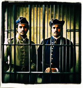 Picture focused on two uniformed men behind bars. Clothes and background appropriate for 16th century France.