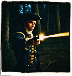 Eighteen year old man firing his flintlock pistol. Located in the dark woods at night, near a dusty road. Scene and clothes appropriate for 16th century France.