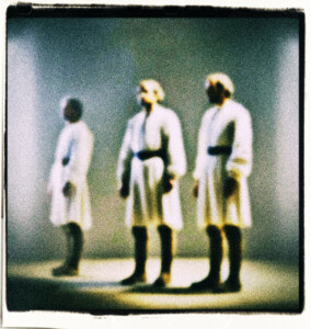 Three copies of the same man looking roughly at the same place. Blurred and bathed in white light, to render them unrecognizable.