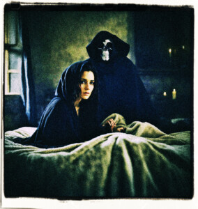 Woman in the bed of a poorly lit room, wearing a black cloak, with the grim reaper in the background.