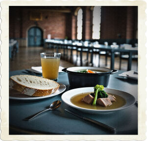 Pictured focused on the food and place setting at the table. there was a sizable piece of bread, a soup composed primarily of broth, and a small plate of steamed meat and vegetables. In contrast to formal dining events, there was one fork, knife, and spoon.  Located in a large dining hall with brick walls and long tables, and sparsely populated.